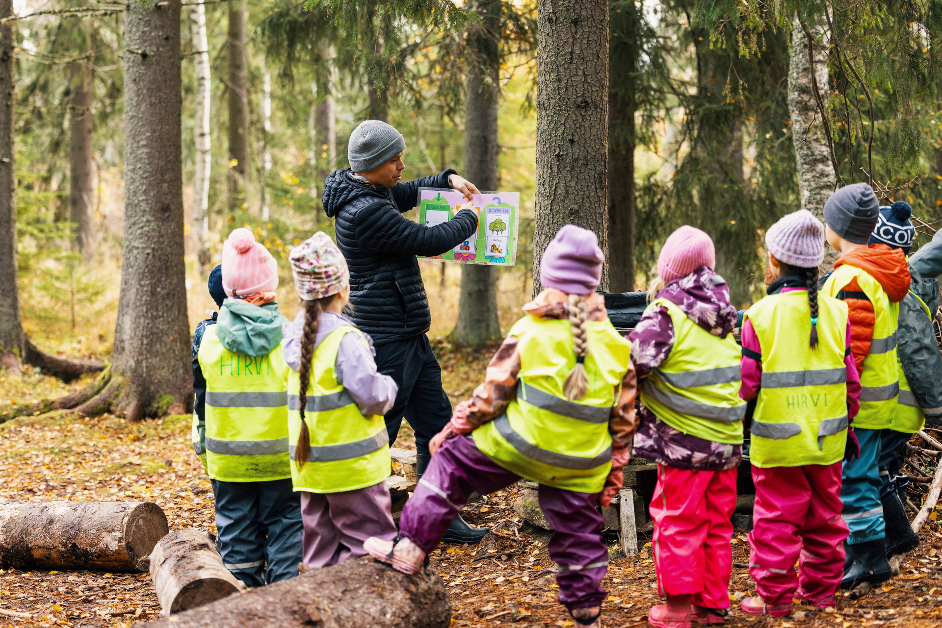 En grupp barn står i en skog och lyssnar på en presentation av en vuxen. Barnen har på sig ljusa reflexvästar och varma ytterkläder.