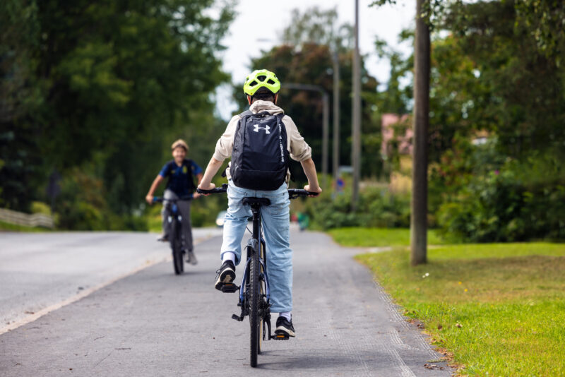 Två barn cyklar på en cykelväg. Barnet i förgrunden har en ljus cykelhjälm och ryggsäck.