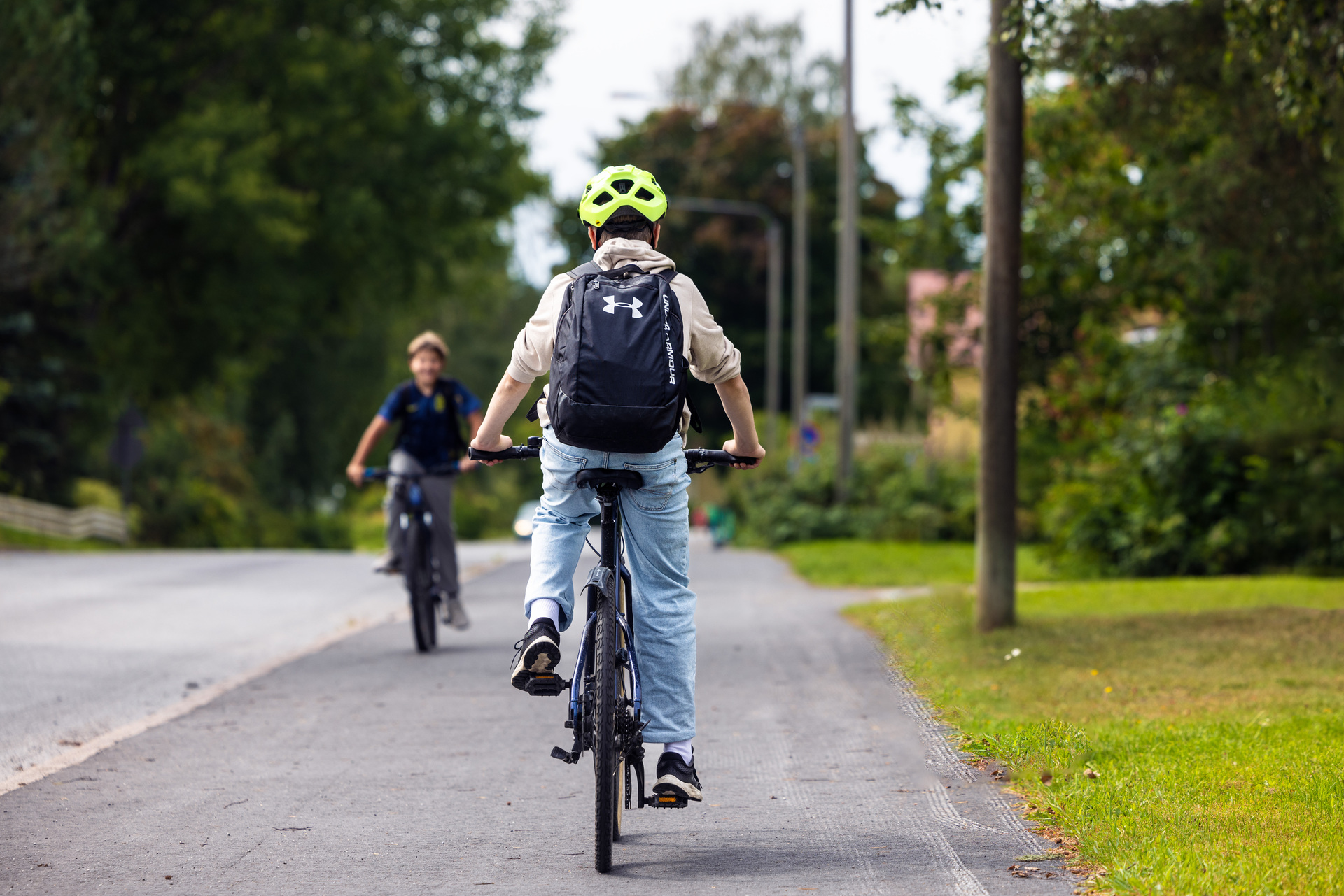 Två barn cyklar på en cykelväg. Barnet i förgrunden har en ljus cykelhjälm och ryggsäck.