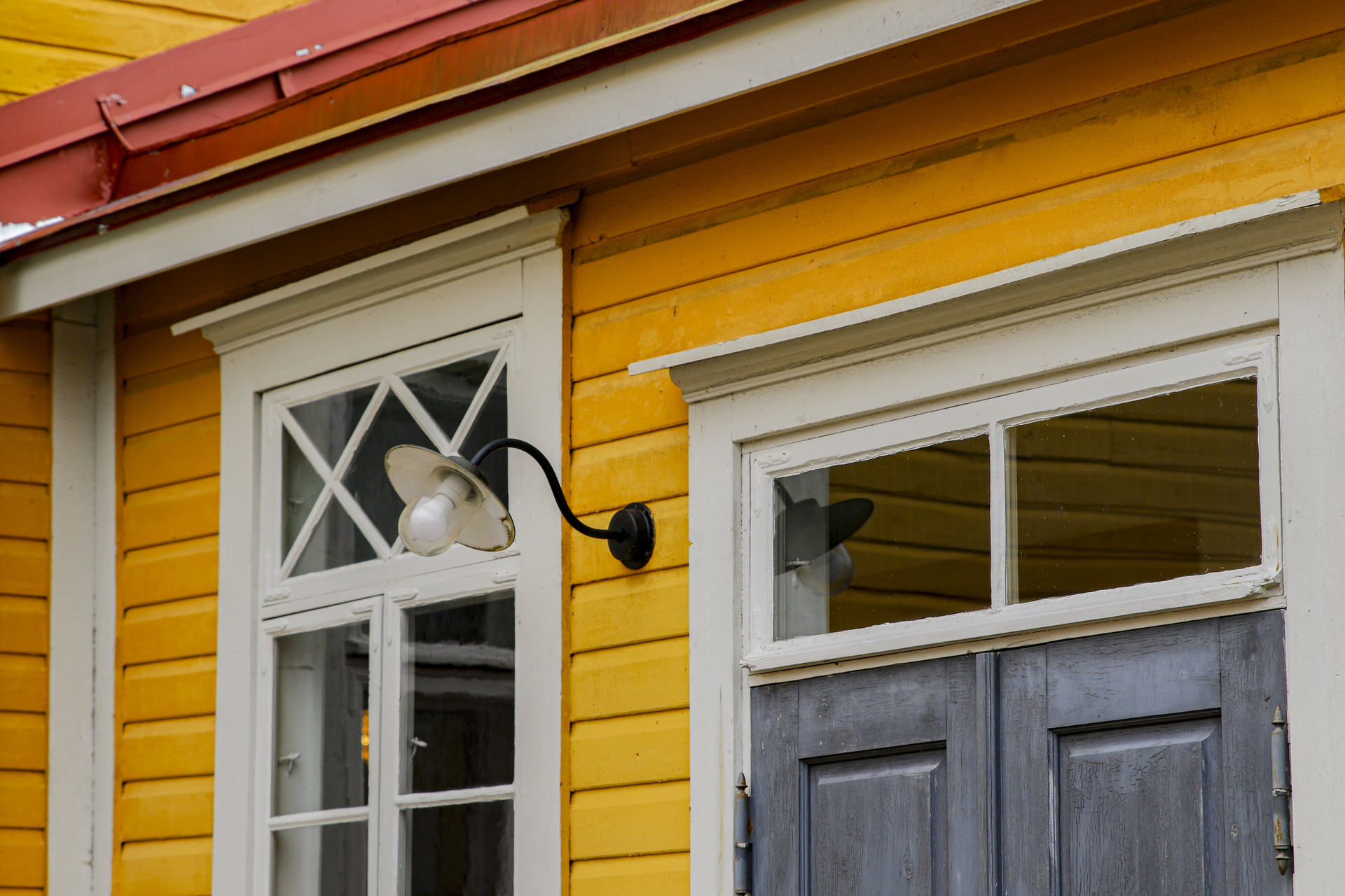 keltaisen puutalon julkisivun lähikuvassa näkyy ikkuna, valaisin ja pääsisäänkäynnin yläosa.