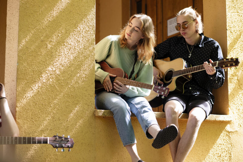 Två personer sitter på en fönsterbräda med gitarrer. Personen till vänster spelar ukulele och personen till höger spelar akustisk gitarr. De är avslappnade och njuter av musiken i starkt solljus.