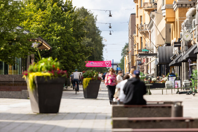 An urban scene with people walking and cycling on a busy street. The street is lined with large flower pots and green trees. In the background, there's a red sign reading "Kävelykatu Sommarågata."