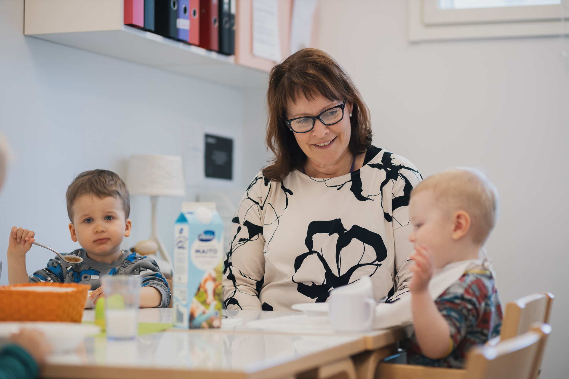 Kaksi lasta syö ruokapöydän ääressä aikuisen seurassa.