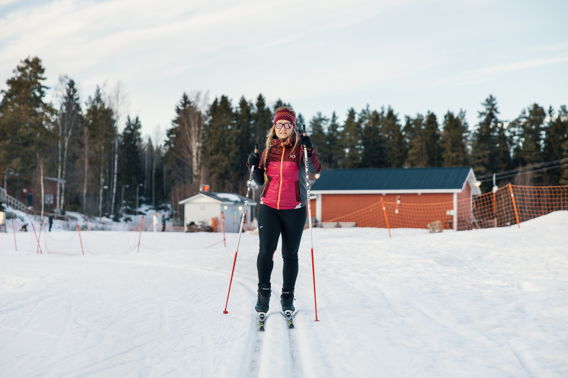 Henkilö hiihtää talvisessa maisemassa punaisen mökin edustalla, taustalla lumisia puita.
