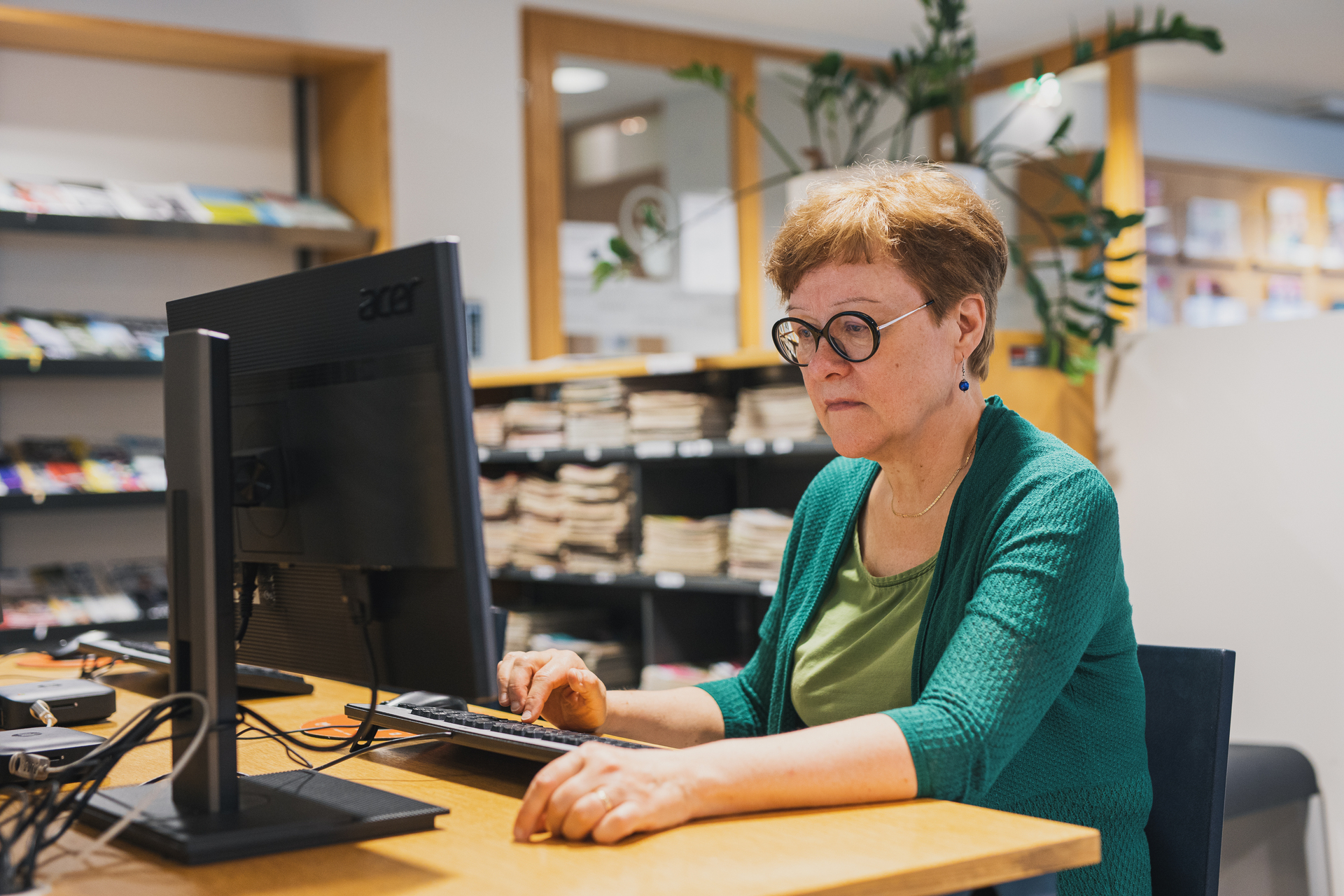 En person som sitter vid en dator på ett bibliotek, med böcker och tidskrifter på hyllor i bakgrunden.