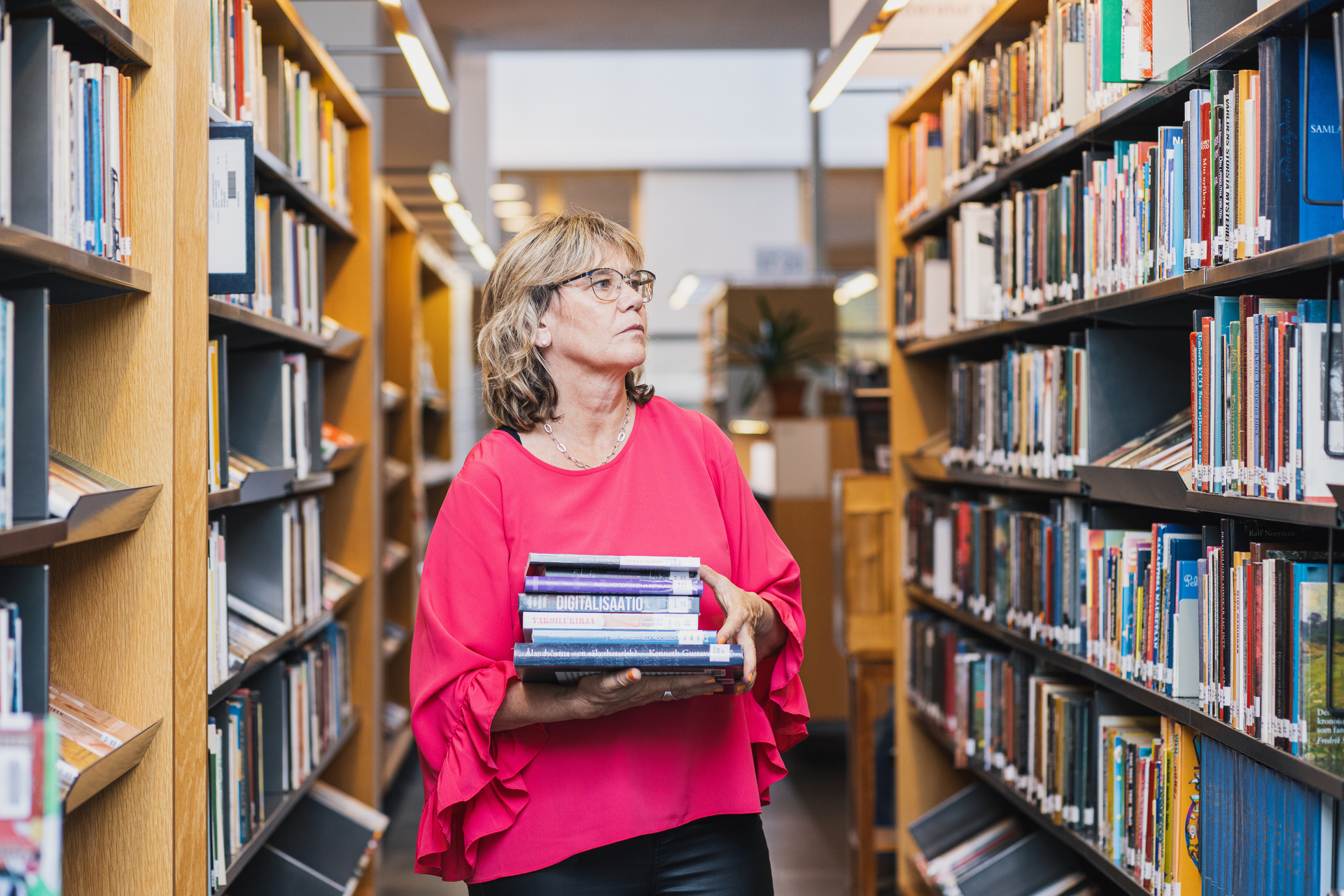 En kvinna med blont hår och rosa tröja bär böcker i ett bibliotek.