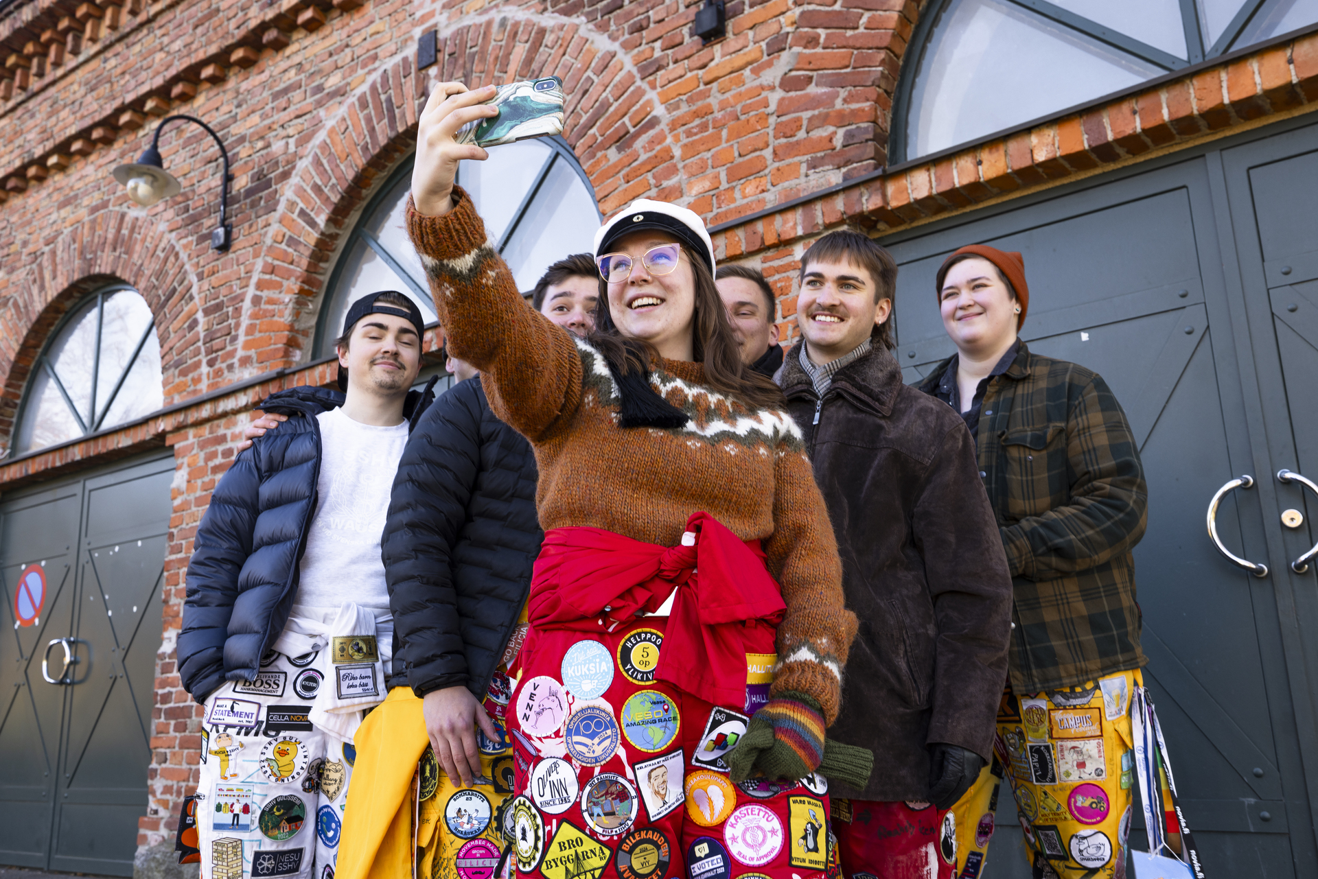 En grupp studenter utanför framför en tegelbyggnad, en av dem tar en selfie. De har på sig studentoveraller med olika märken.