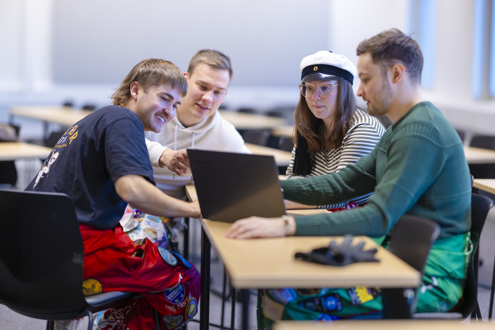 Neljä opiskelijaa istuu luokkahuoneessa pöydän ääressä, katsellen kannettavaa tietokonetta. Heillä on yllään värikkäät opiskelijahaalarit ja yhdellä on valkoinen teekkarilakki.