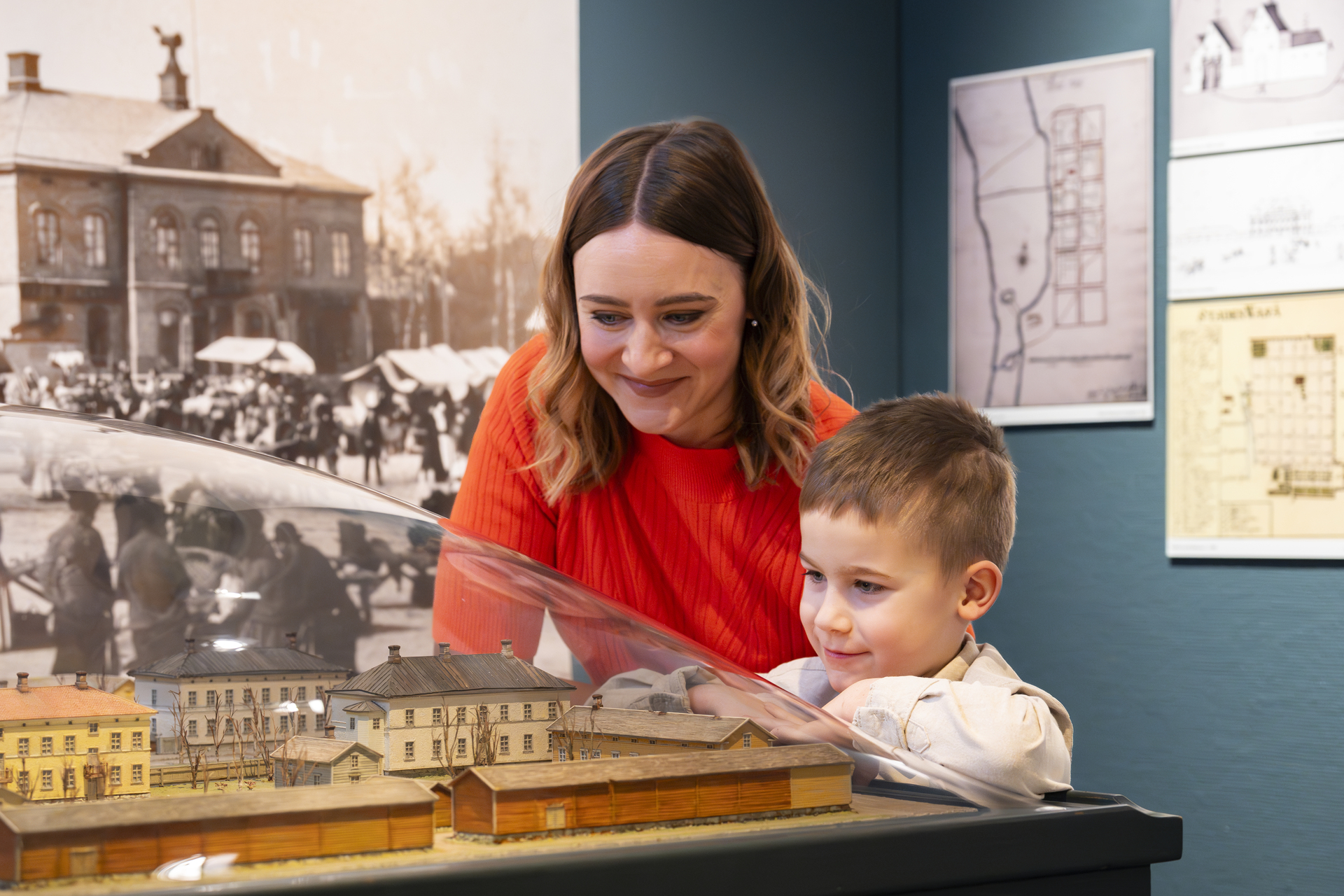 A woman and a child look at a miniature model in a museum with historical photographs in the background.