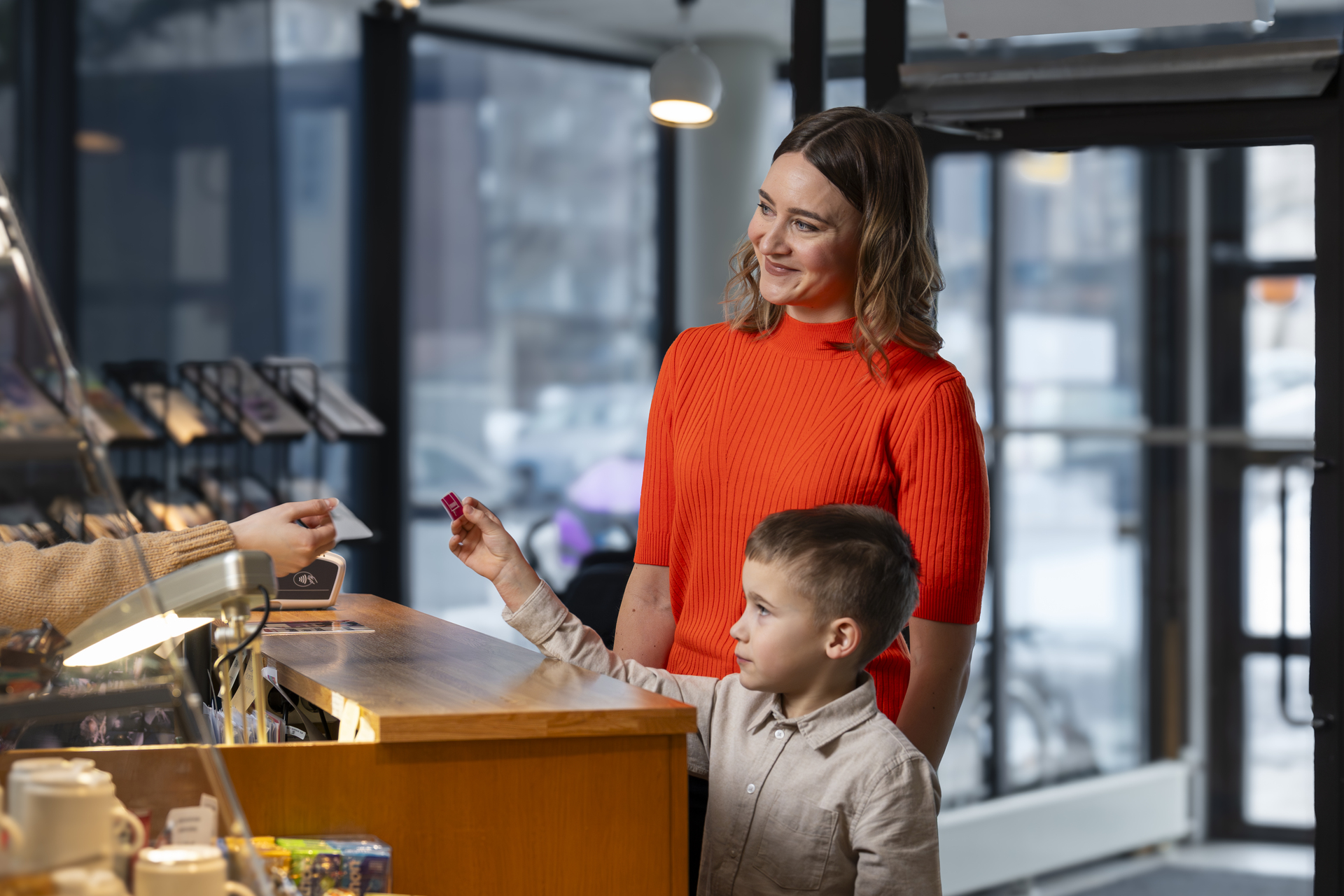 A woman and child at a counter in a café, the woman is smiling and the child is handing something to the cashier.