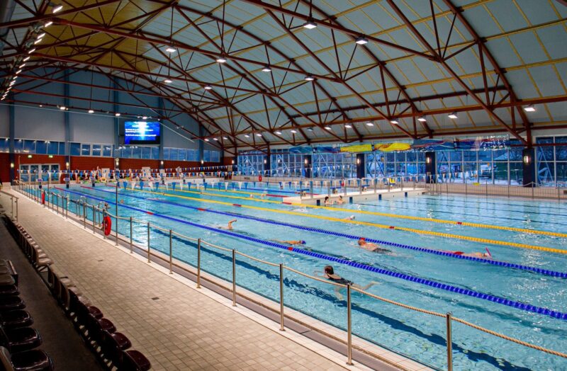Inomhuspool med flera banor. Människor simmar och åskådare sitter på läktarna.