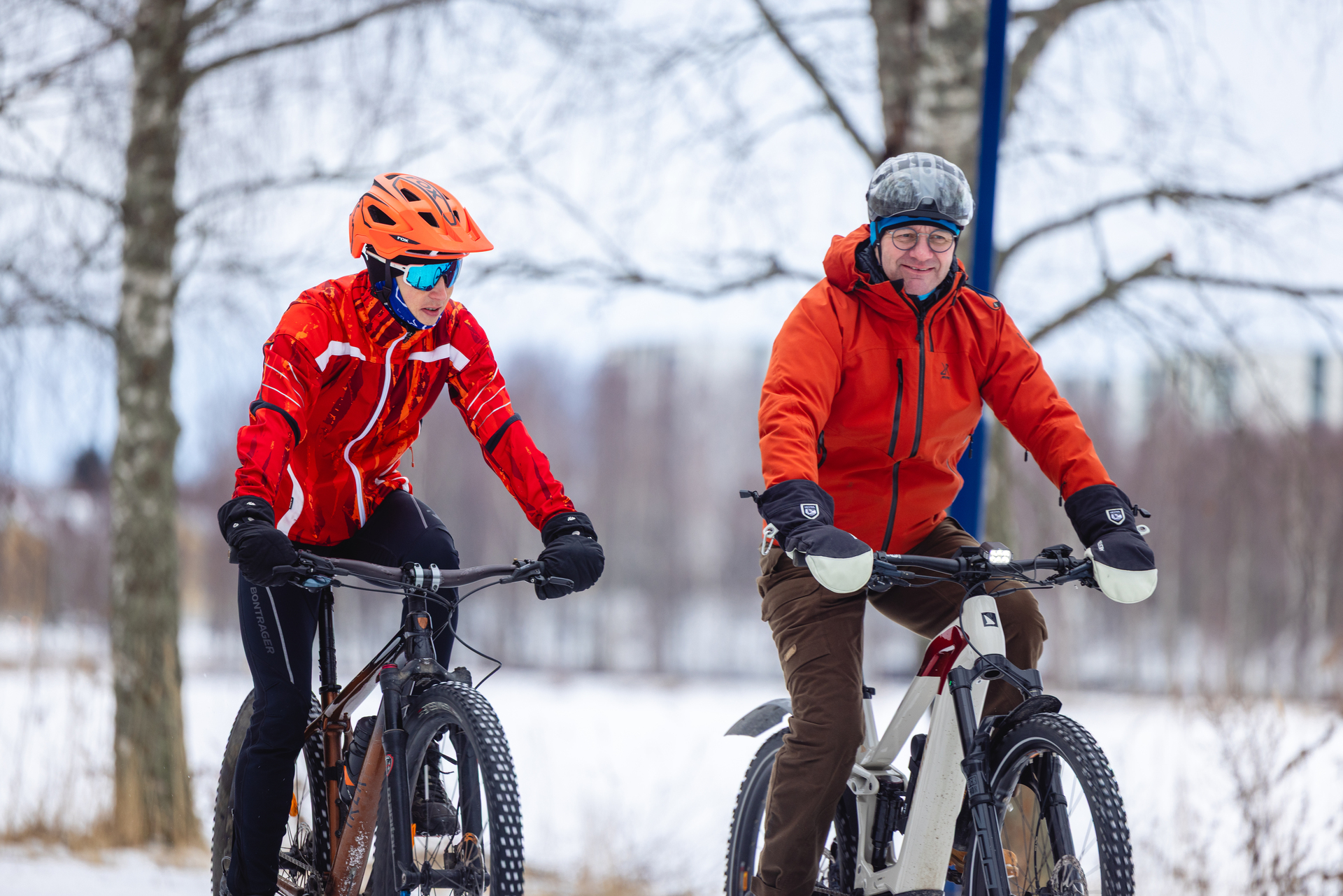 Två personer cyklar i ett vinterlandskap, klädda i orange jackor och hjälmar.