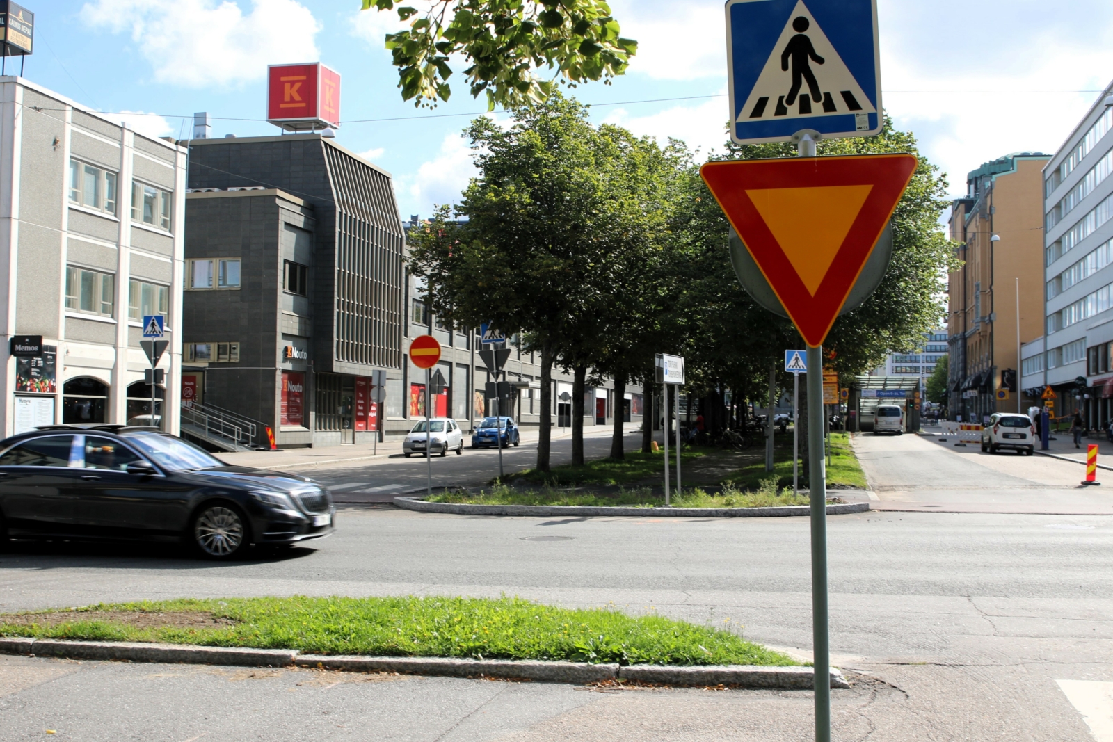 Kaupunkimaisema, jossa on suojatien merkki ja väistämisvelvollisuusmerkki. Taustalla on liikkuvia autoja ja rakennuksia.