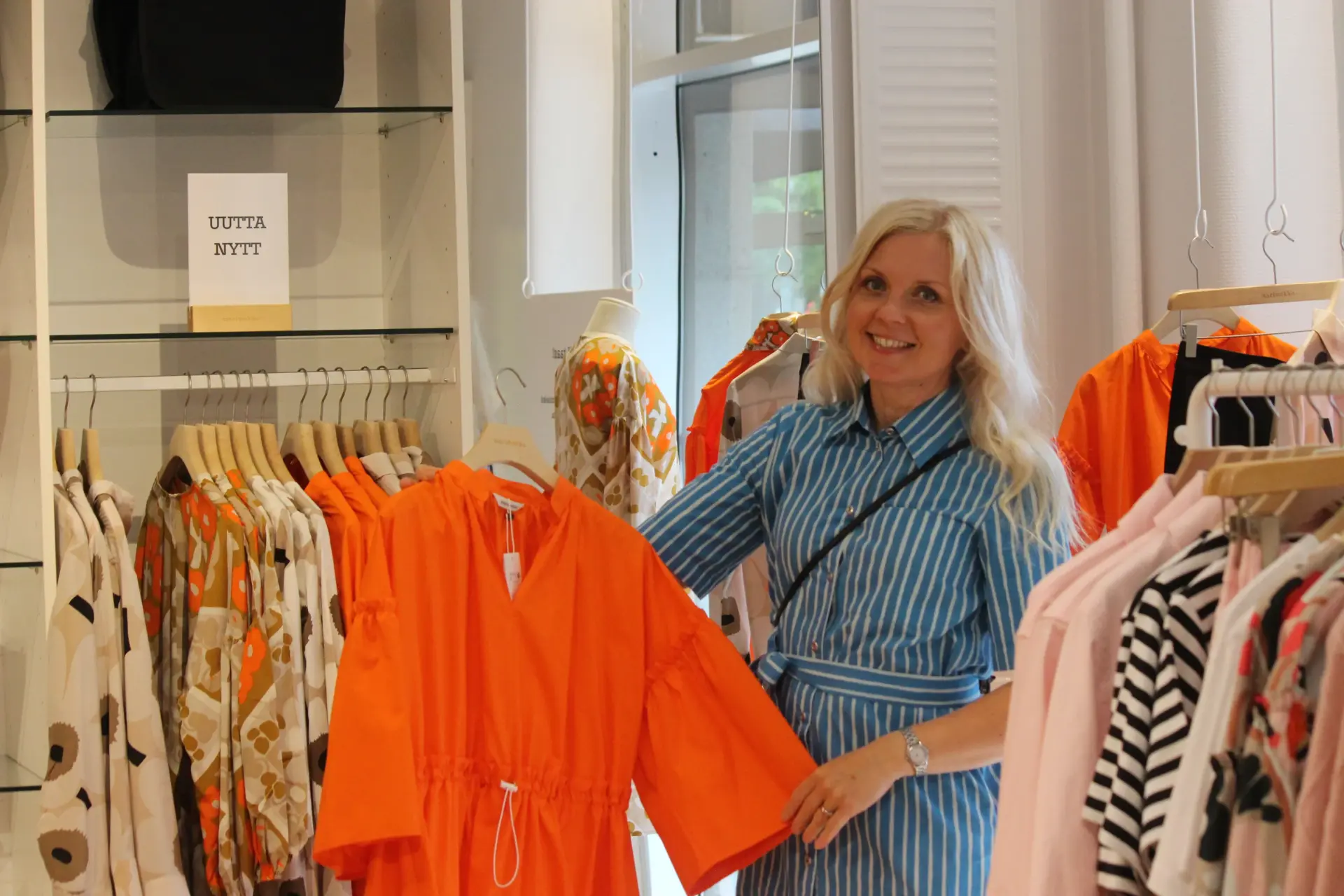 A woman smiles in a shop and displays an orange garment on a rack, with other colorful clothes in the background.