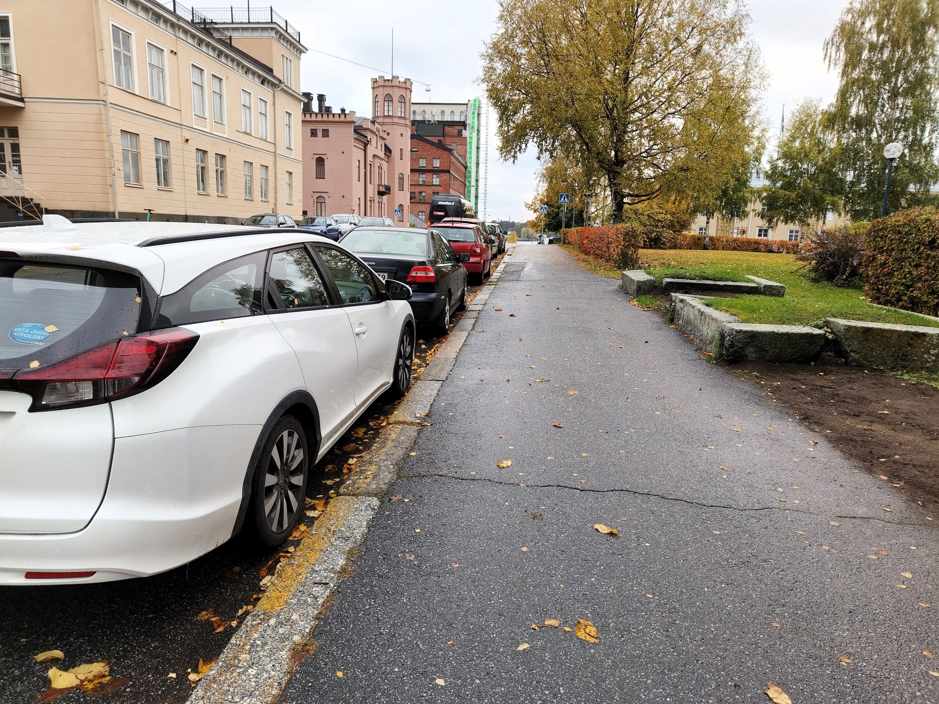 Pysäköityjä autoja kadun varrella syksyllä, taustalla vaaleanpunainen rakennus ja puita.
