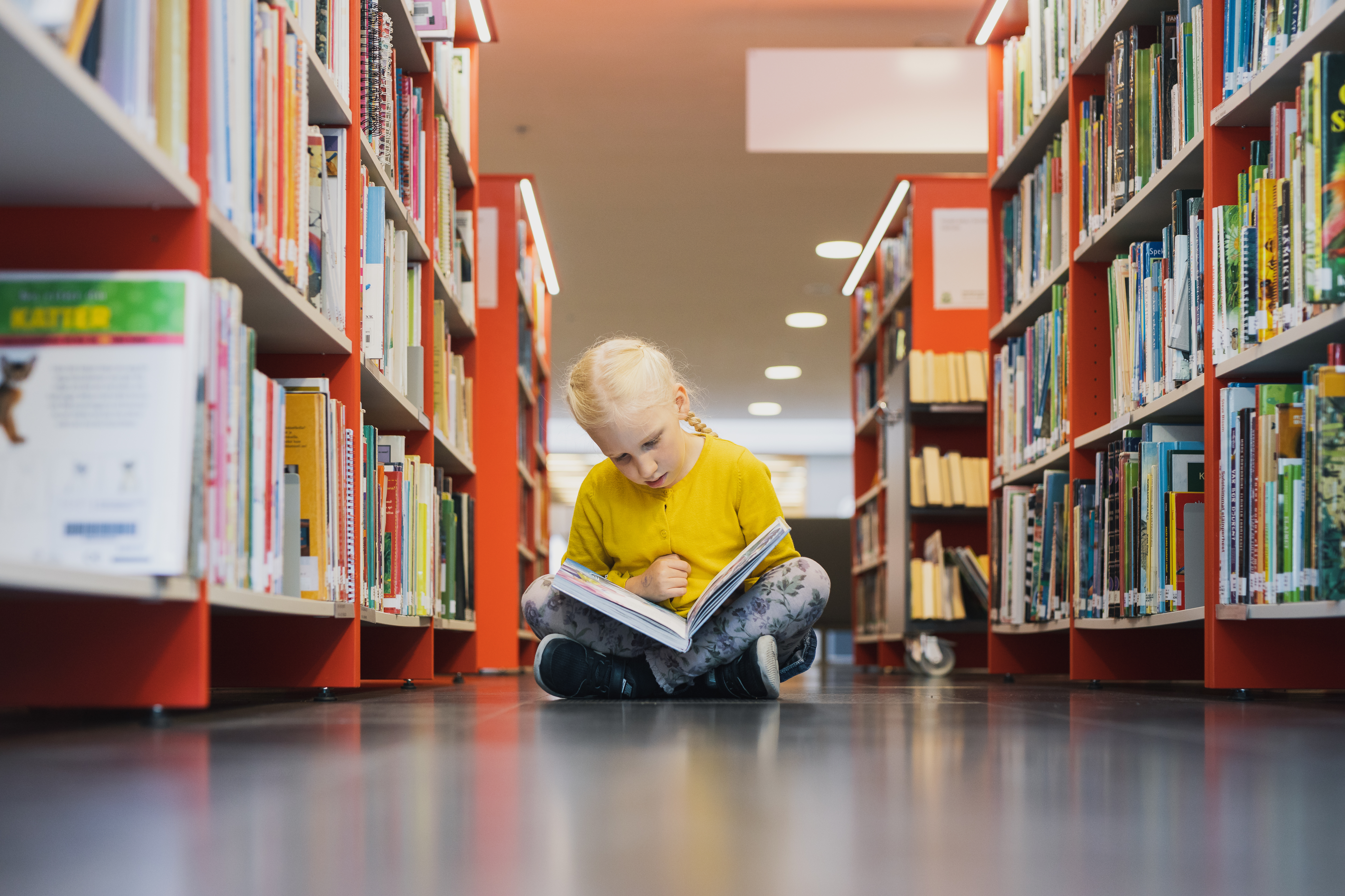 A girl sitting on the floor of a library reading a book.