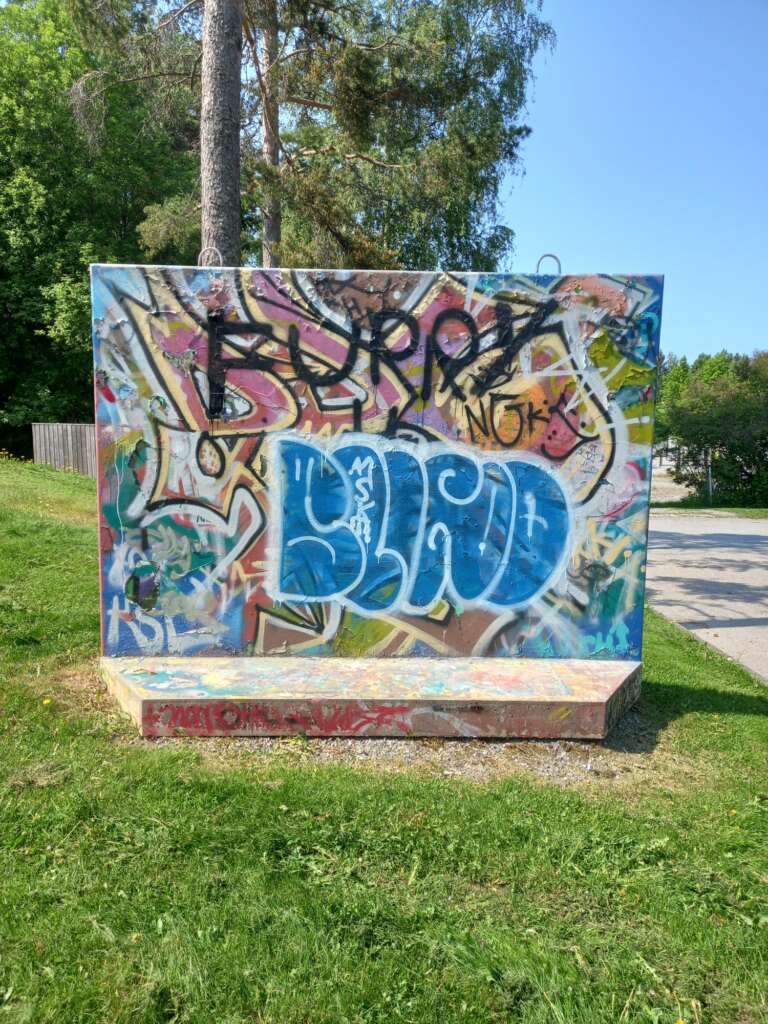 Graffitivägg med färgglada målningar och skrifter i en grön park.