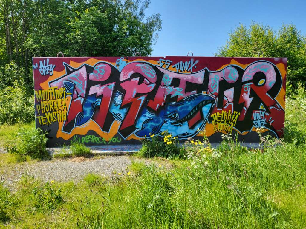 Graffitivägg med en färgglad målning. Texten lyder "Muuttori työskalee Texasitin" och "Delikki Autti". I mitten finns en blå djurfigur som påminner om en val. Grön växtlighet i bakgrunden.