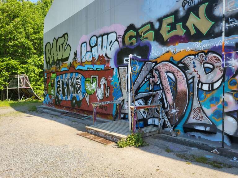En vägg med färgglad graffitikonst med stora bokstäver och geometriska former, samt en liten metallramp på marken.