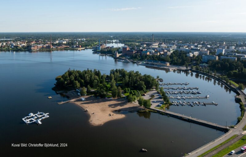 Flygfoto av en strand och småbåtshamn med en liten skogsklädd ö, stad i bakgrunden.