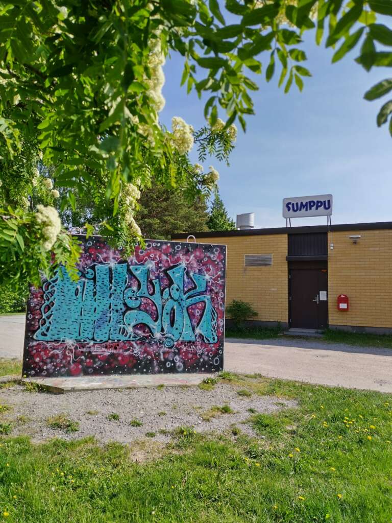 Graffitikonst på en utomhusvägg med texten "SUMPPU". Det växer frodiga träd i närheten och solen skiner starkt.