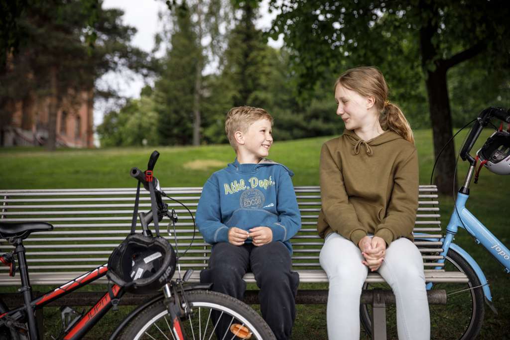 Kaksi lasta istuu penkillä puistossa hymyillen toisilleen. Heidän vieressään on kaksi polkupyörää.