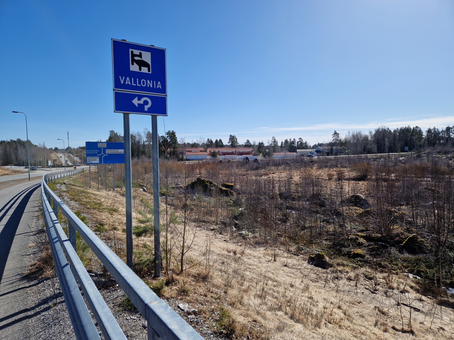 Bilden visar en väg med en vägskylt som pekar mot Vallonia. I bakgrunden finns träd och byggnader. Det är en solig dag.