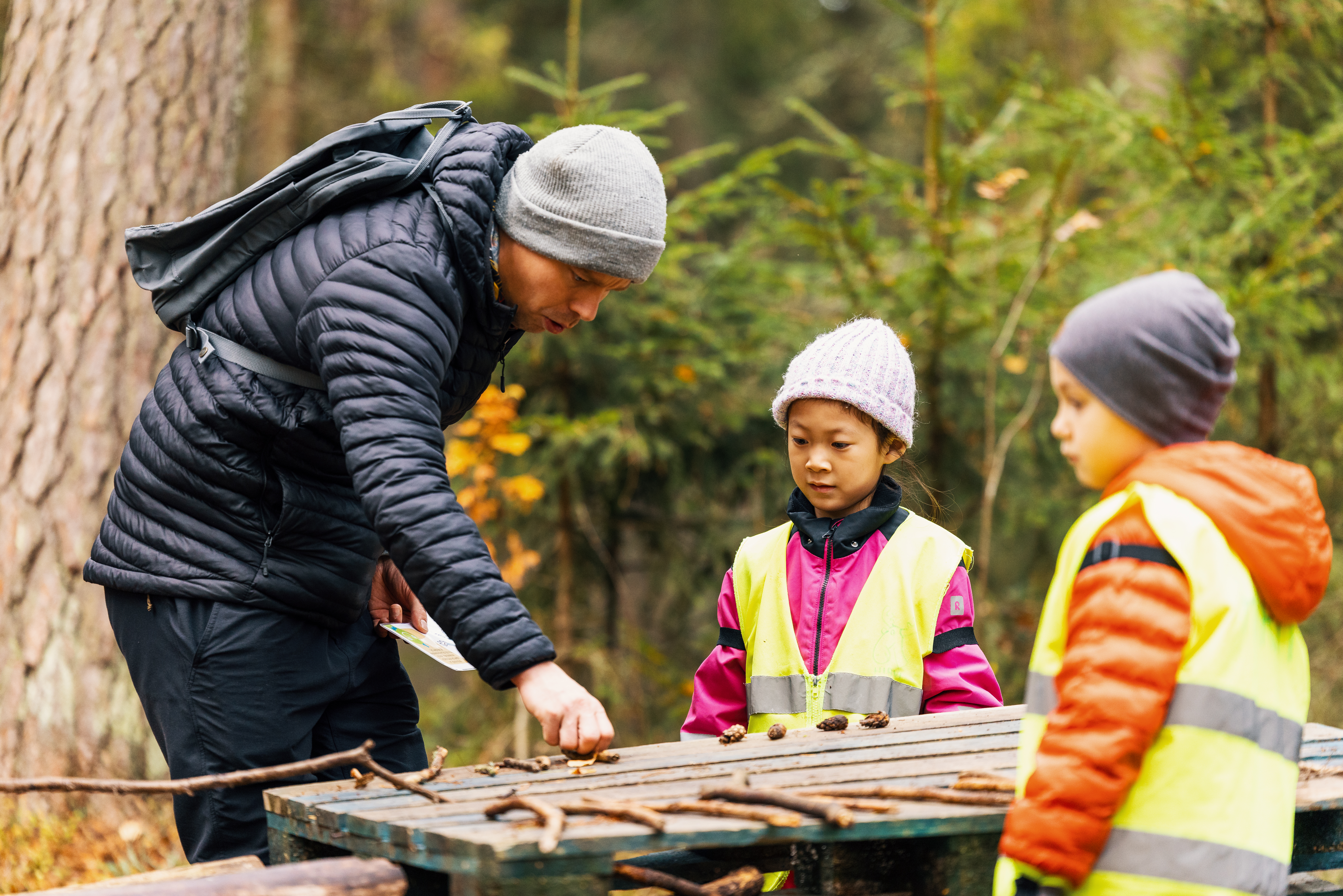 En vuxen och två barn ute i skogen. Den vuxne visar barnen pinnar och stenar som är arrangerade på ett utomhusbord. Barnen har på sig ljusa säkerhetsvästar och mössor.