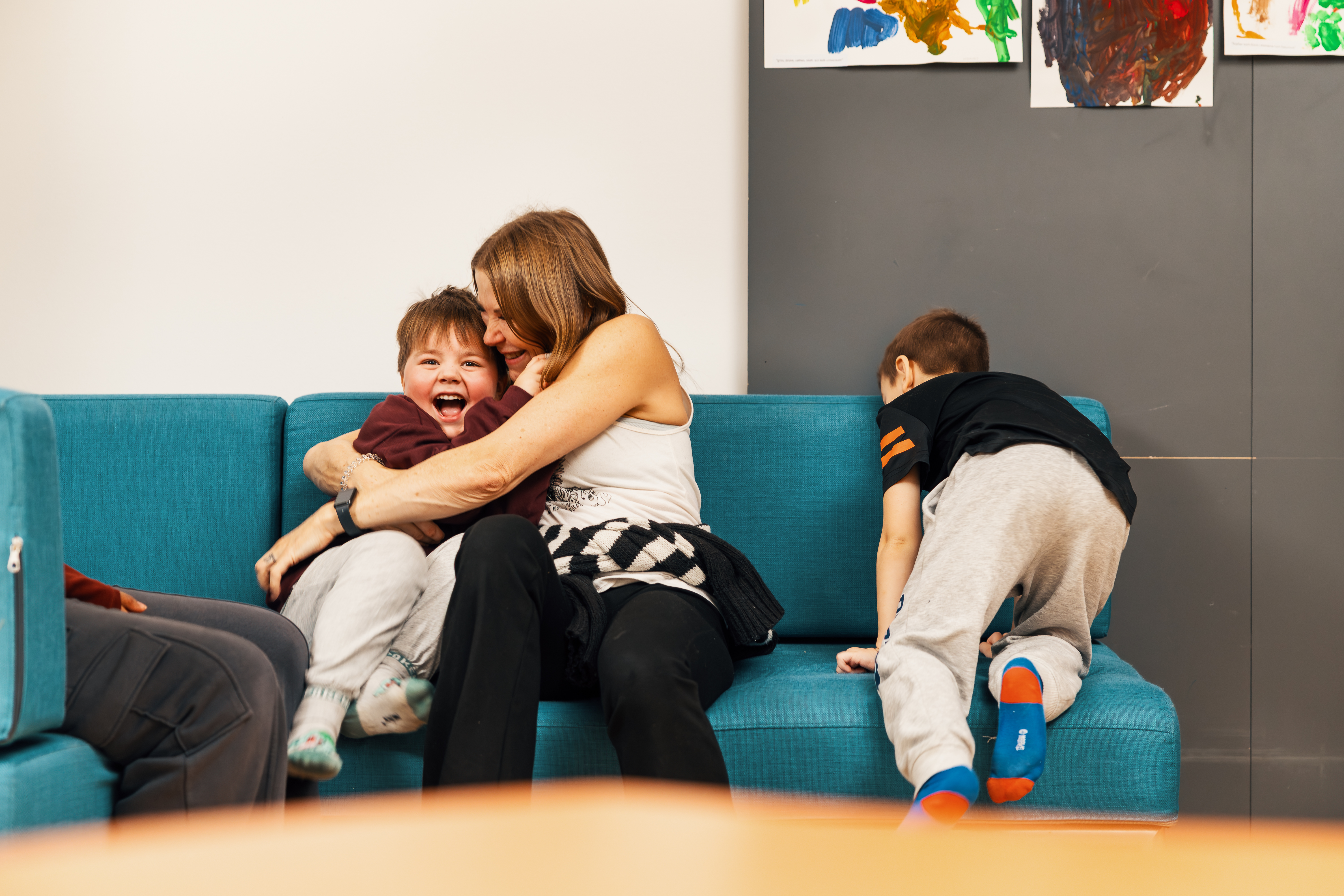 A woman hugging a child on a couch, the child is laughing happily. Another child is playing on the other side of the couch.