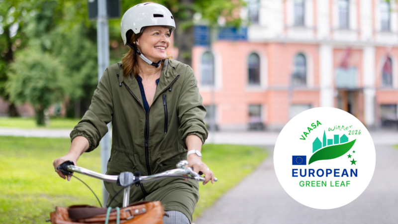 En kvinna cyklar i en park med en byggnad i bakgrunden. Bilden inkluderar också Vasa European Green Leaf 2026-vinnarsymbolen.