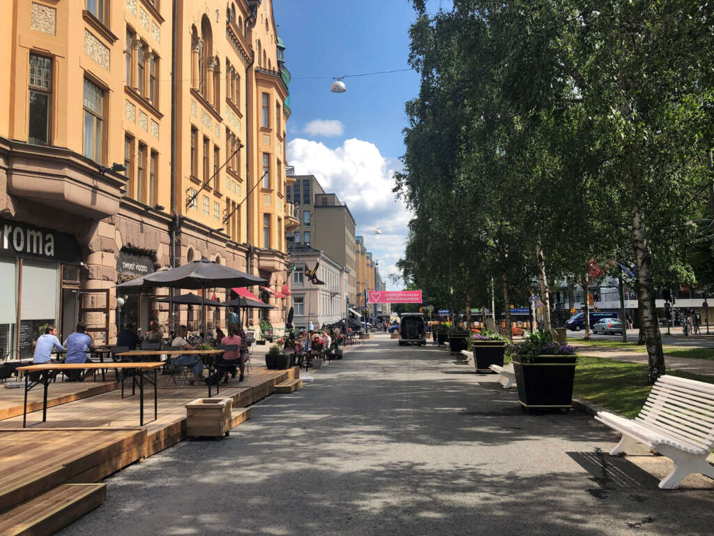 En gågata kantad med kaféer och blomlådor på en solig dag. Människor sitter utomhus på terrasser. Det finns en gammal byggnad till vänster och träd till höger.