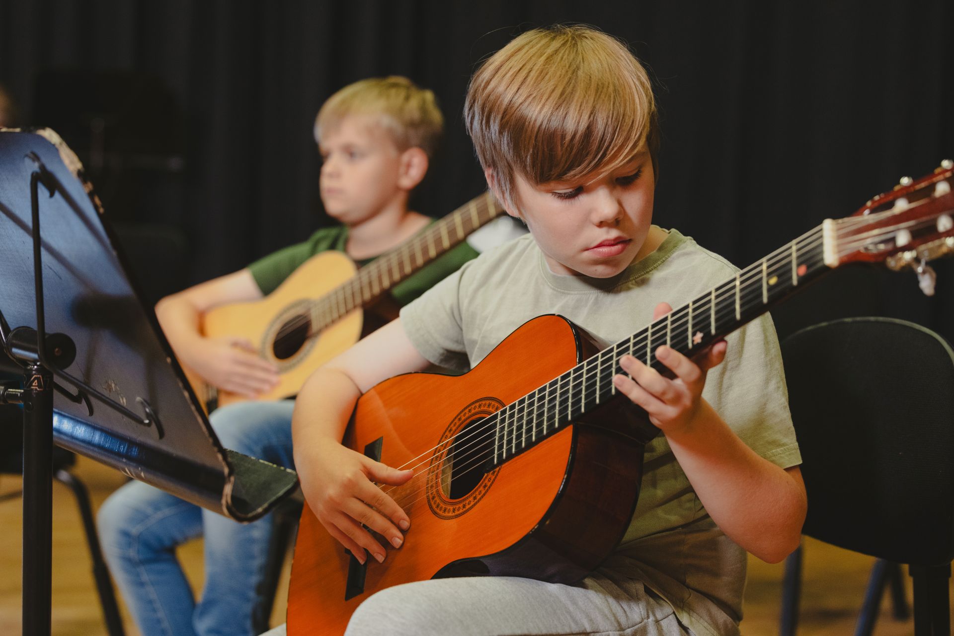 Two young boys playing acoustic guitars while sitting on chairs during a music lesson.