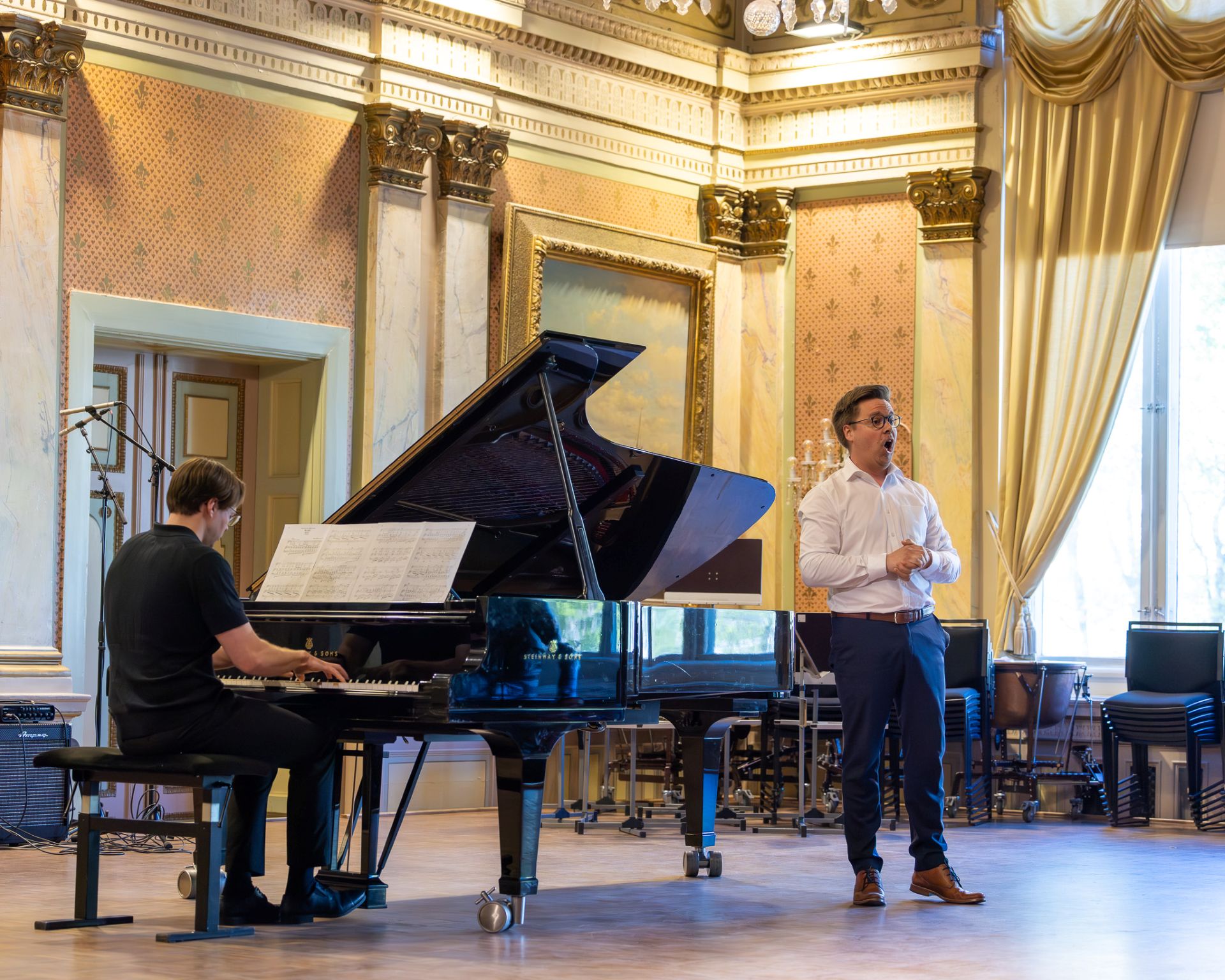 Bild av en lyxig konserthall med guldförgyllda väggar och stora fönster. Till vänster sitter en man och spelar på en flygel, och till höger står en sångare i vit skjorta och blå byxor och sjunger.