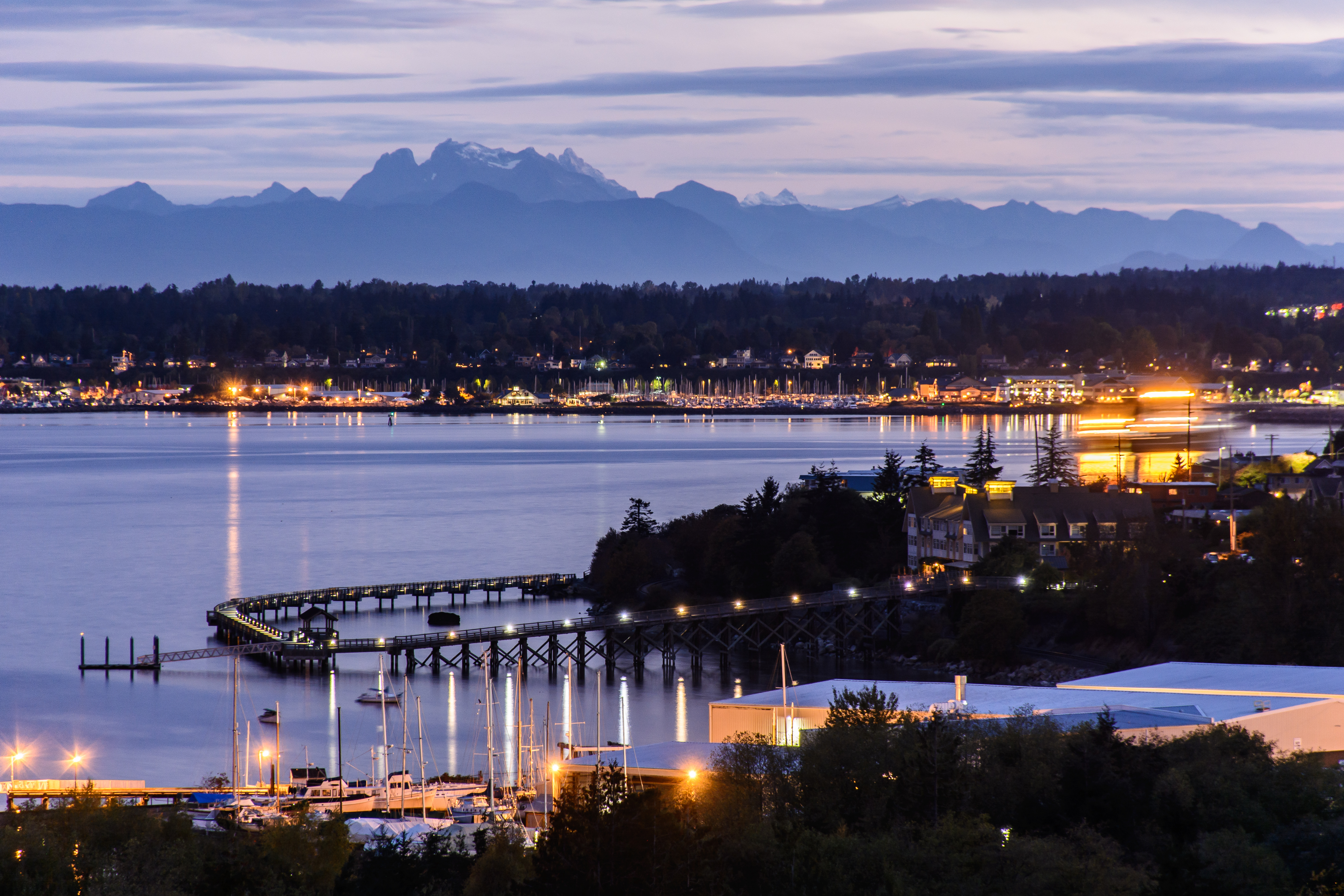 A scenic evening view of a coastal city with shimmering lights reflecting on the water. In the background, mountains are silhouetted against the twilight sky.