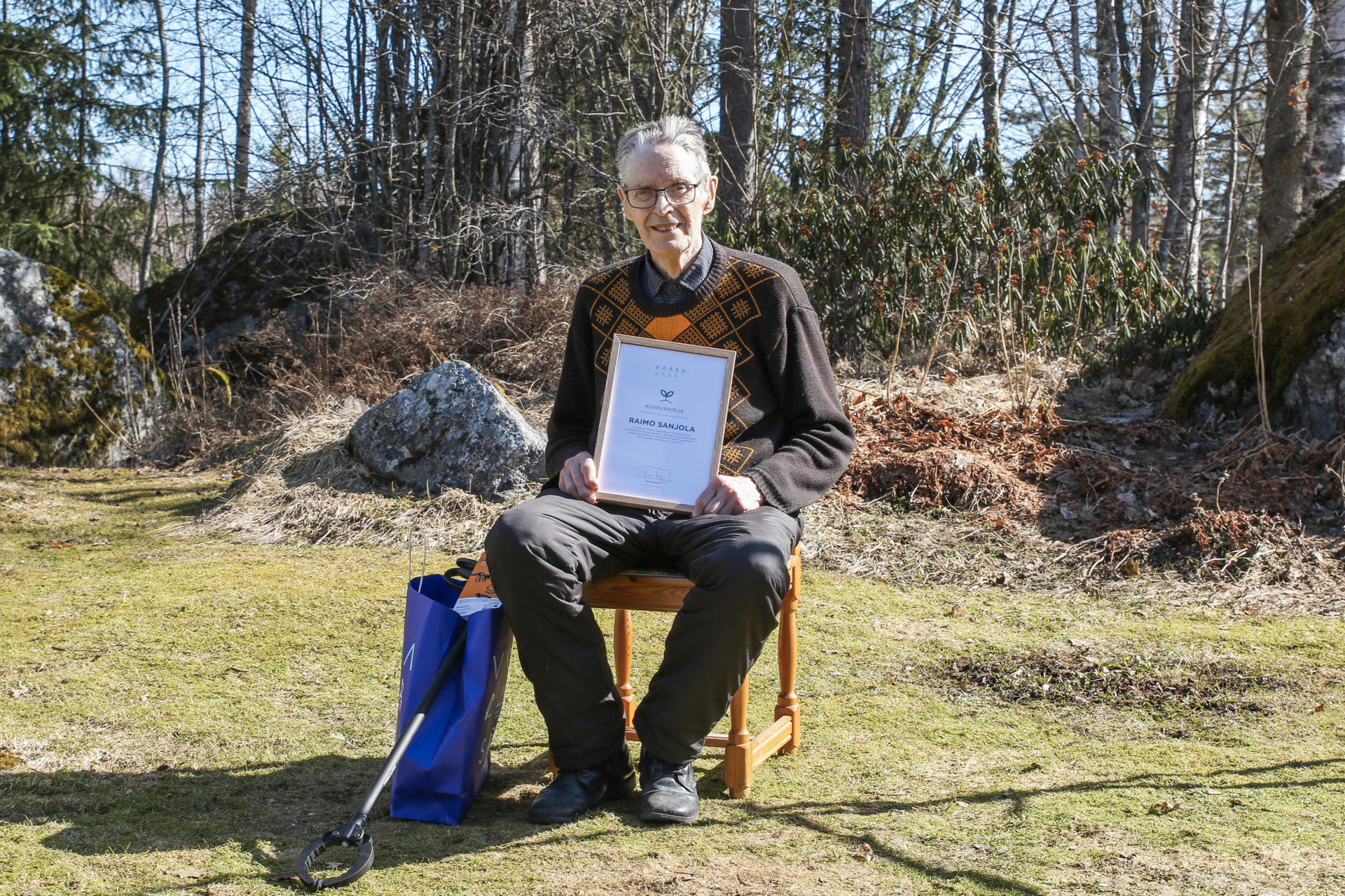 Henkilö istuu puutarhassa tuolilla sylissään kehystetty diplomi. Aurinko paistaa, taustalla näkyy puita.