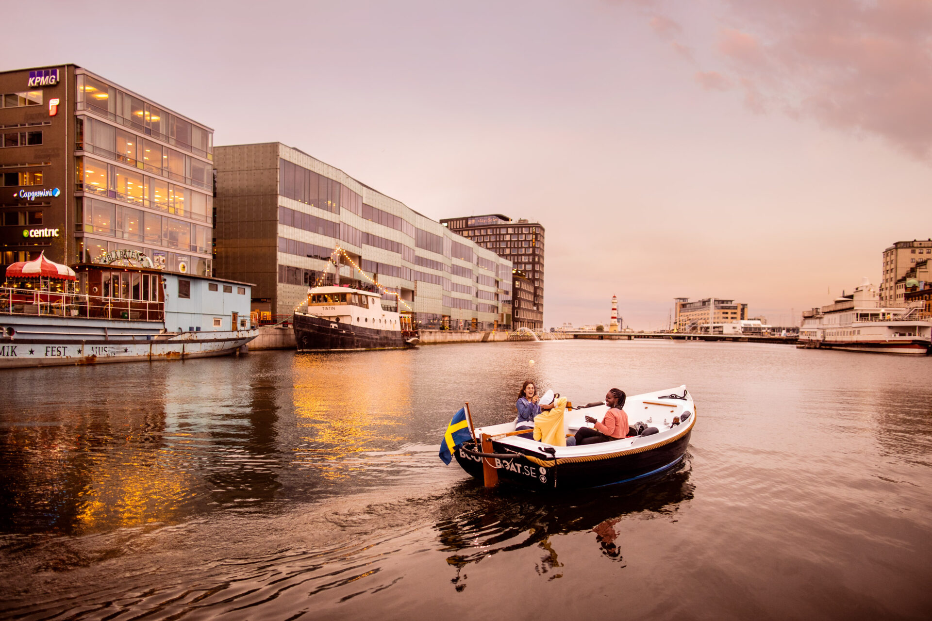A boat cruising in Malmö canal next to a modern building, with several boats and a cityscape in the background.