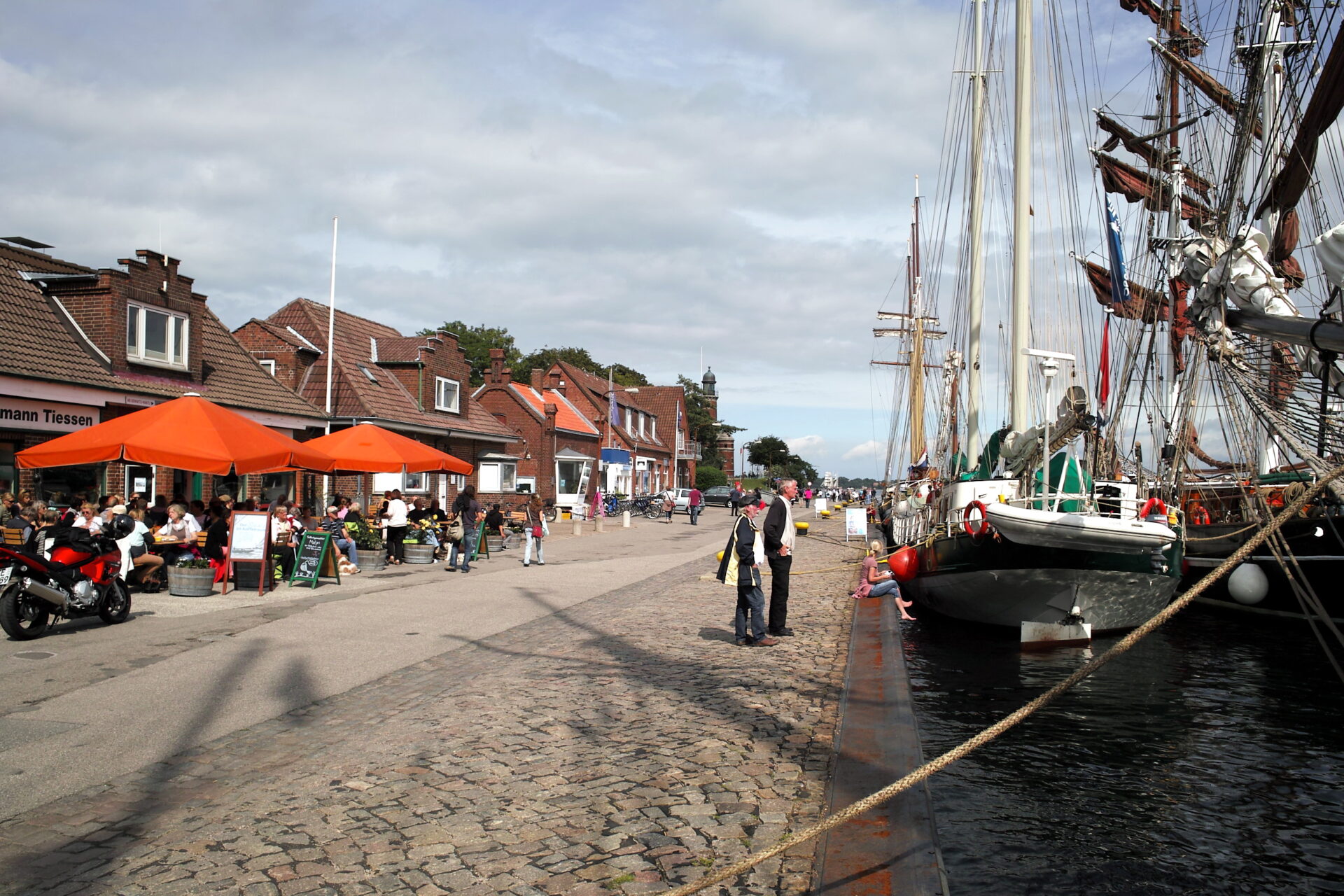 Seaside street with a restaurant, large sailing ships, and people walking.