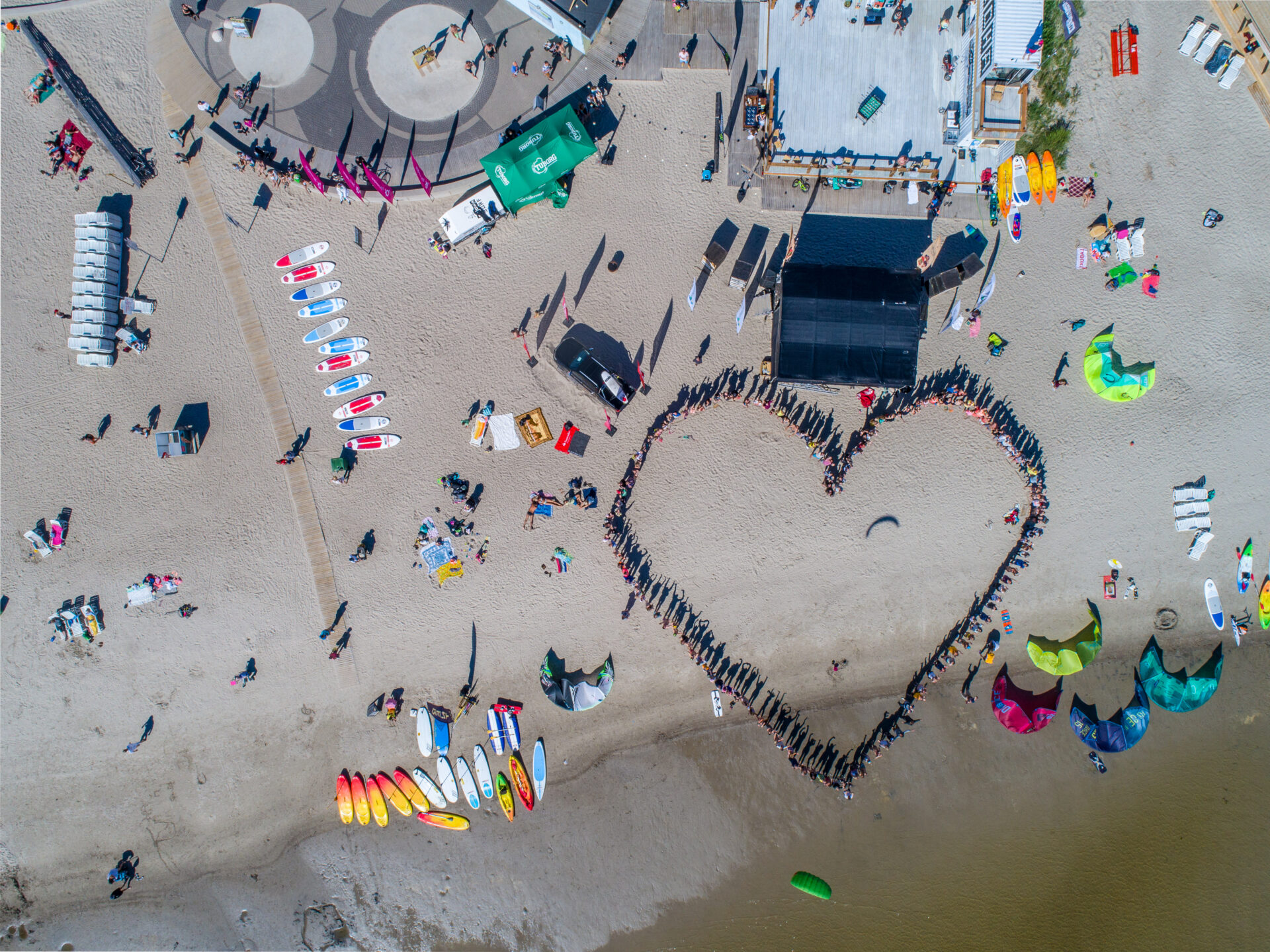 Aerial view of a beach where people have formed a heart shape on the sand. The beach also shows colorful surfboards and equipment.