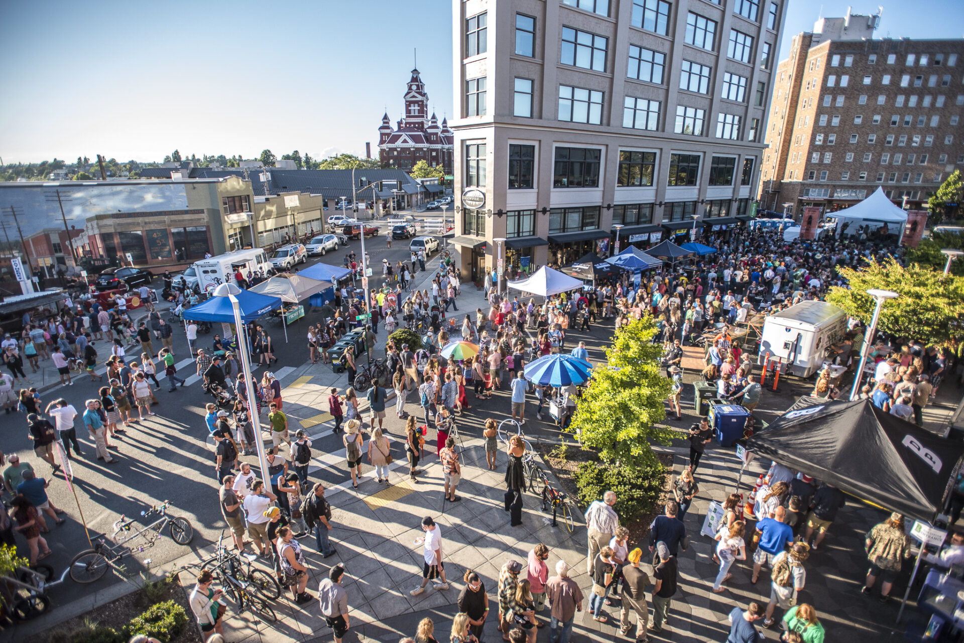 A crowd of people attending an outdoor event in a city center on a bright day. There are canopy tents and stalls on the surrounding streets.
