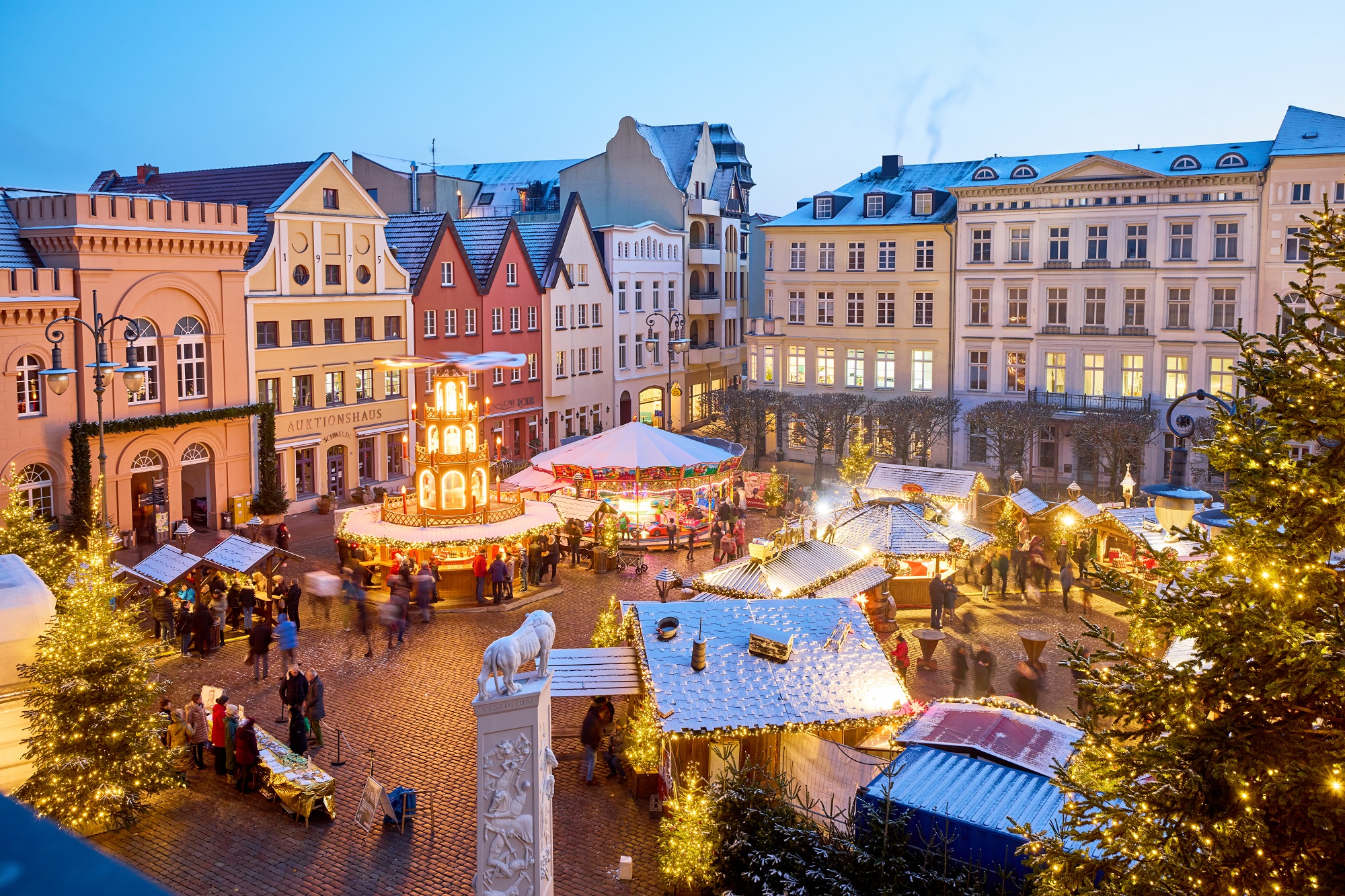 En julmarknad på ett gammalt torg, dekorerat med ljusslingor och julgranar, med människor som strosar runt stånden.