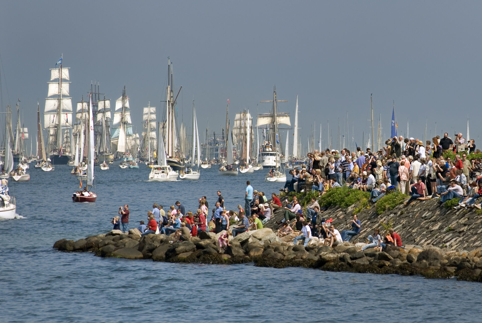 People sitting on rocks watching sailing ships on the sea.