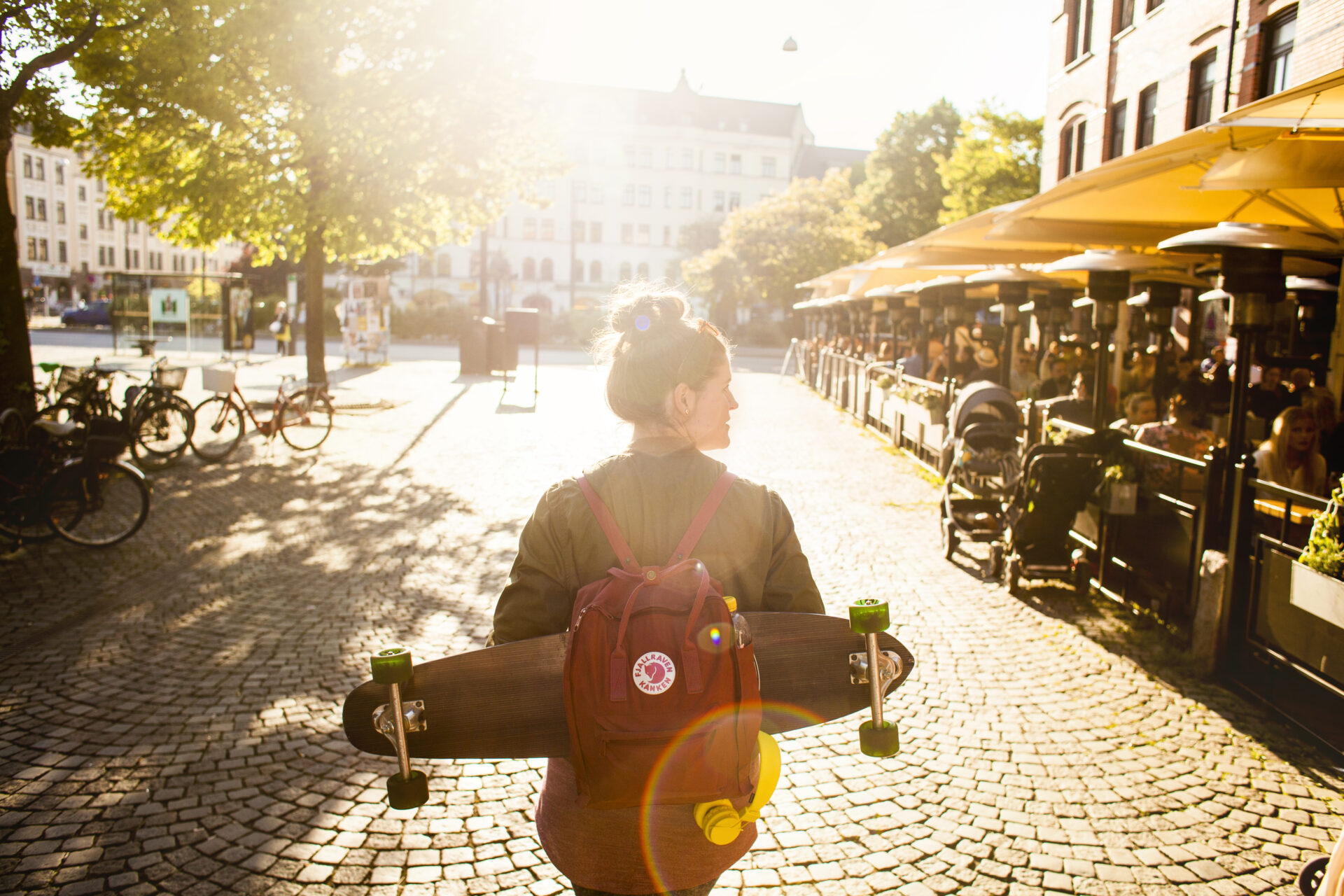 A person carrying a skateboard on a Malmö street with cafes and bicycles on one side.