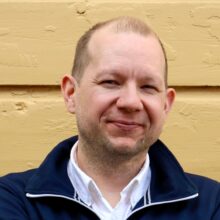 A man standing in front of a yellow wall, looking at the camera. He has short light hair and is wearing a dark blue jacket and a white shirt.