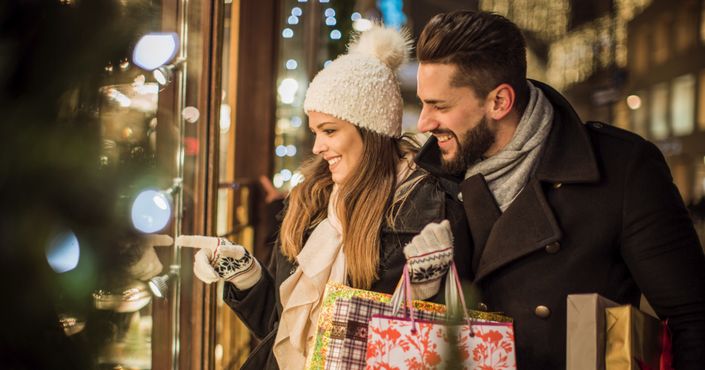 Two people looking at a festively decorated shop window on a shopping street lit up for Christmas, holding shopping bags.