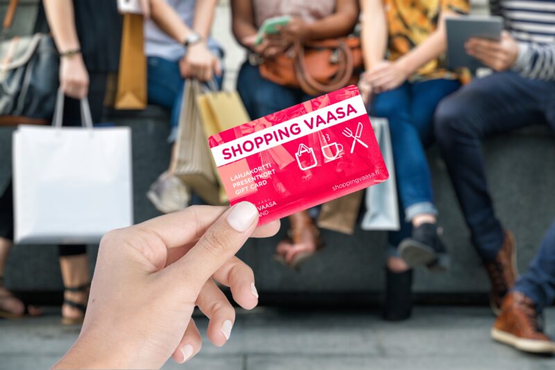 A person holding a Shopping Vaasa gift card with people sitting on a bench with shopping bags in the background.