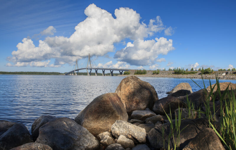 Bilden visar en stor bro som sträcker sig över vatten, med stenar i förgrunden på stranden. Det finns stora vita moln på himlen.