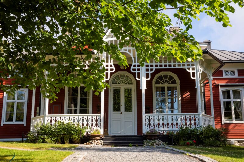 Kaunis punainen puutalo, jossa on valkoinen koristeellinen kuisti ja vehreä piha.