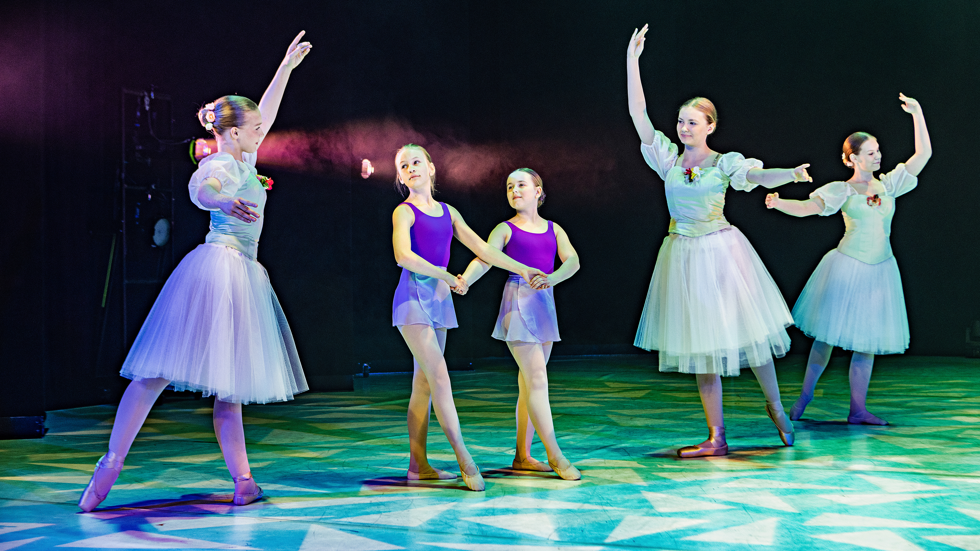 Young dancers in ballet poses under colorful stage lighting.