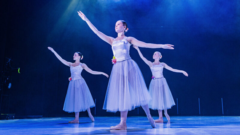 Three ballet dancers perform a choreography on stage under blue lighting. They are all wearing white tutus with floral decorations at the waist.