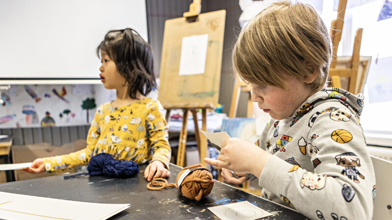 Two children crafting at a table. The girl on the left looks away while holding a ball of yarn, and the boy on the right is cutting paper with scissors. Drawings and an easel are visible in the background.